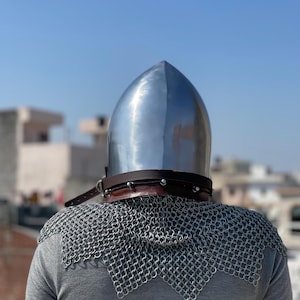 May include: A shiny metal helmet with a pointed crown, a brown leather neck guard, and a chainmail collar. The helmet is worn over a grey long-sleeved top. The background shows a clear blue sky and buildings.