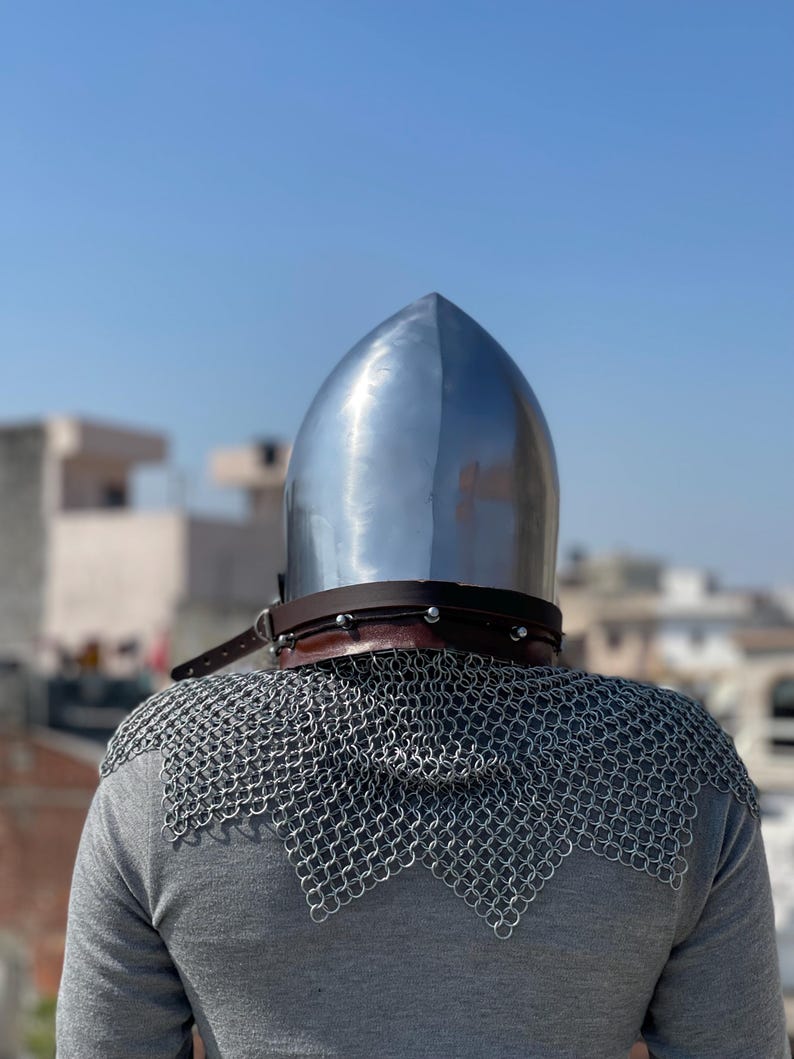 May include: A shiny metal helmet with a pointed crown, a brown leather neck guard, and a chainmail collar. The helmet is worn over a grey long-sleeved top. The background shows a clear blue sky and buildings.