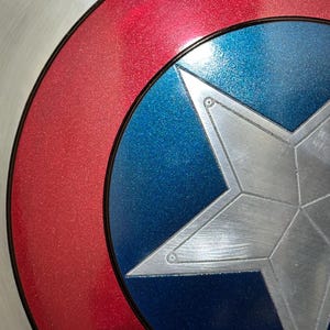 May include: Close-up of a Captain America shield. The shield features a silver star on a blue background, surrounded by a red ring and a silver outer ring. The shield has a metallic finish.