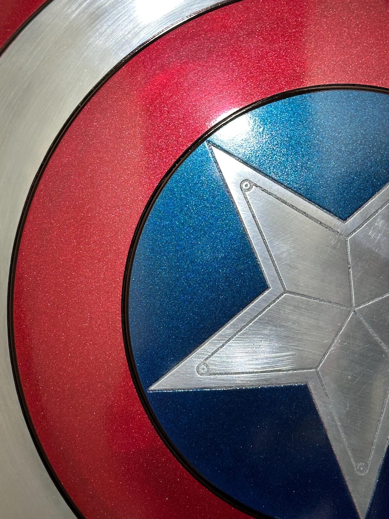May include: Close-up of a Captain America shield. The shield features a silver star on a blue background, surrounded by a red ring and a silver outer ring. The shield has a metallic finish.