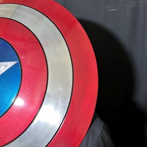 May include: A red, white, and blue shield with a star emblem. The shield has concentric circles in red, silver, and white. The center features a blue field with a white star.