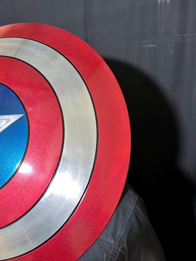 May include: A red, white, and blue shield with a star emblem. The shield has concentric circles in red, silver, and white. The center features a blue field with a white star.