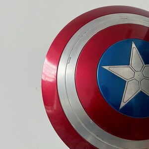May include: A red, white, and blue shield with a silver star in the center. The shield has a circular design with concentric rings of red, silver, and blue. The shield is a prop or collectible.