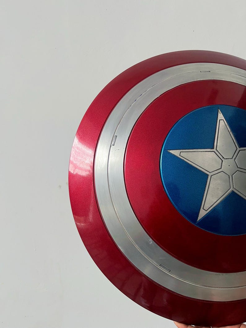 May include: A red, white, and blue shield with a silver star in the center. The shield has a circular design with concentric rings of red, silver, and blue. The shield is a prop or collectible.