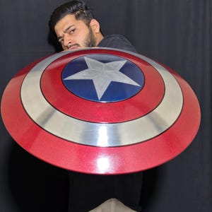 May include: A red, white, and blue shield with a star emblem. The shield has a circular design with a silver band separating the red and blue sections. A classic superhero accessory.