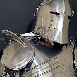 May include: A polished steel knight's helmet and breastplate. The helmet features eye slits, a cross design, and decorative metalwork. The breastplate and shoulder armor are riveted together. Chainmail is visible at the neck.