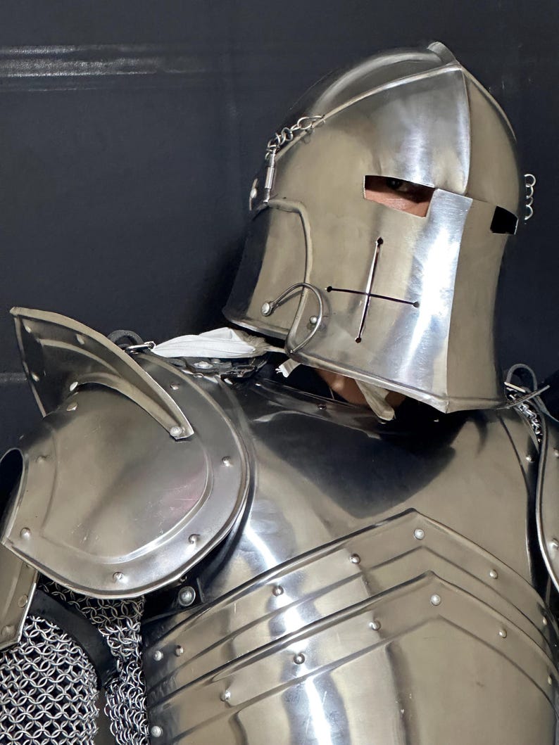 May include: A polished steel knight's helmet and breastplate. The helmet features eye slits, a cross design, and decorative metalwork. The breastplate and shoulder armor are riveted together. Chainmail is visible at the neck.