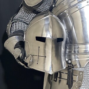 May include: A polished steel knight's helmet with a cross design, held against a backdrop of matching armor. The helmet features eye slits and a rounded top. The armor includes chainmail and articulated plates, showcasing medieval craftsmanship.