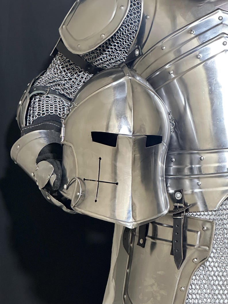 May include: A polished steel knight's helmet with a cross design, held against a backdrop of matching armor. The helmet features eye slits and a rounded top. The armor includes chainmail and articulated plates, showcasing medieval craftsmanship.
