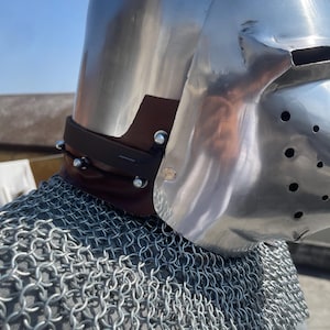 May include: A shiny metal helmet with a visor, brown leather neck guard, and a chainmail shirt. The helmet reflects the light and is secured with rivets. The chainmail is made of interlocking metal rings.