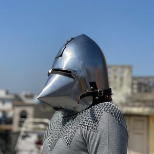 May include: A shiny steel medieval helmet with a pointed visor and a leather strap. The helmet is worn over a grey shirt and chainmail armour. The background is a clear blue sky and buildings.