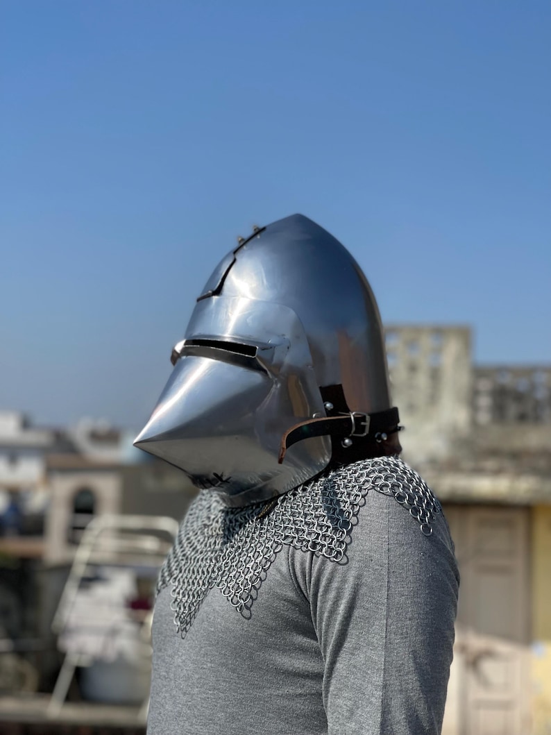 May include: A shiny steel medieval helmet with a pointed visor and a leather strap. The helmet is worn over a grey shirt and chainmail armour. The background is a clear blue sky and buildings.