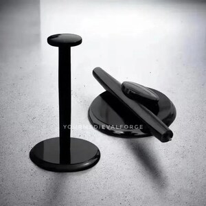 May include: Two black display stands. One is tall with a circular base and a flat, round top. The other is low, with a circular base and a curved, cylindrical top. The stands are on a light grey surface.