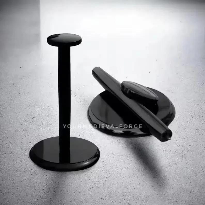 May include: Two black display stands. One is tall with a circular base and a flat, round top. The other is low, with a circular base and a curved, cylindrical top. The stands are on a light grey surface.