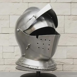 May include: A polished, silver-colored knight's helmet with a visor and neck guard. The helmet has a series of small holes near the face and is mounted on a black stand. The background is a light-colored brick wall.