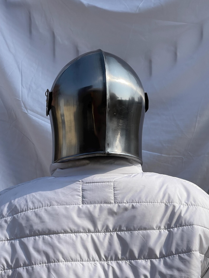 Peut inclure: Un casque en acier poli avec un sommet arrondi et une visi&egrave;re incurv&eacute;e. Le casque est r&eacute;fl&eacute;chissant, montrant l'environnement. Fait partie d'une armure m&eacute;di&eacute;vale.