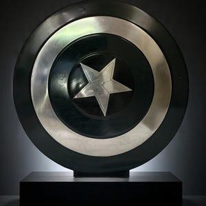 Puede incluir: Un escudo negro y plateado con un diseño de estrella plateada en el centro. El escudo tiene forma circular con un centro negro, un anillo plateado y un borde exterior negro. Se muestra sobre una base rectangular negra.