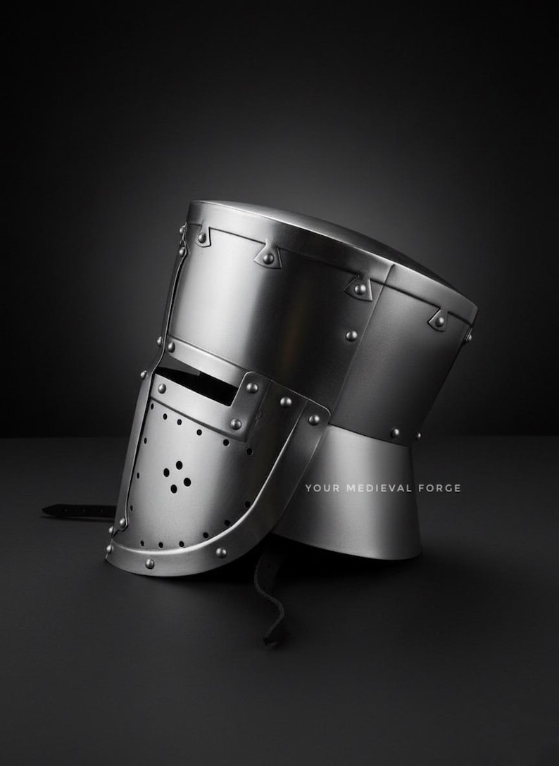 Puede incluir: Un casco medieval pulido de color plateado con una construcci&oacute;n remachada. El casco presenta una visera con m&uacute;ltiples agujeros peque&ntilde;os y una protecci&oacute;n para el cuello. El texto "YOUR MEDIEVAL FORGE" est&aacute; impreso en la parte inferior derecha.