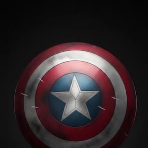 May include: A round shield with a red, white, and blue color scheme. The shield features a white star on a blue circle in the center, surrounded by concentric red and white bands. The shield has a distressed appearance.