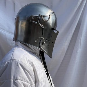 Peut inclure: Un casque de chevalier en acier poli avec une visi&egrave;re orn&eacute;e d'une croix. Le casque a un dessus arrondi et une visi&egrave;re articul&eacute;e, reli&eacute;e par une cha&icirc;ne. Le casque fait partie d'une armure m&eacute;di&eacute;vale.