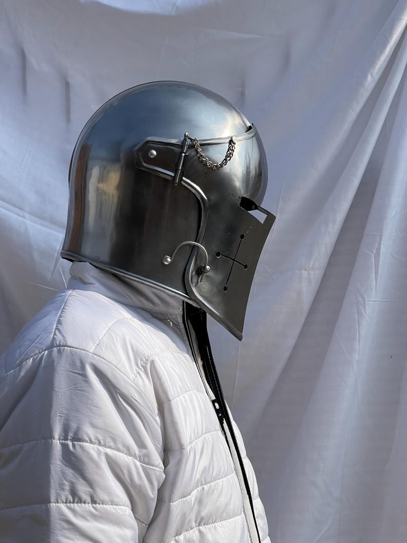 Peut inclure: Un casque de chevalier en acier poli avec une visi&egrave;re orn&eacute;e d'une croix. Le casque a un dessus arrondi et une visi&egrave;re articul&eacute;e, reli&eacute;e par une cha&icirc;ne. Le casque fait partie d'une armure m&eacute;di&eacute;vale.
