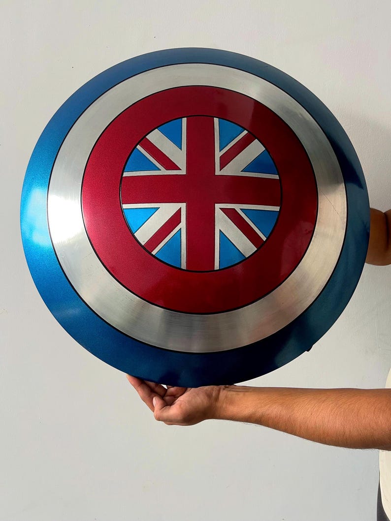May include: A round shield with a blue outer ring, a silver band, and a red center featuring a Union Jack design. The shield is held up against a white background.