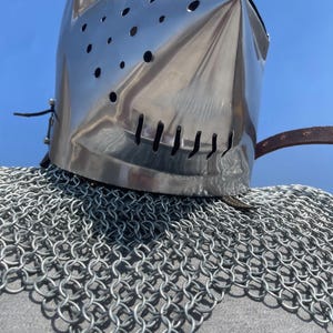 May include: A shiny, silver knight's helmet with a pointed visor and ventilation holes. The helmet rests on a chainmail coif, with a leather strap. The background is a clear, bright blue sky.