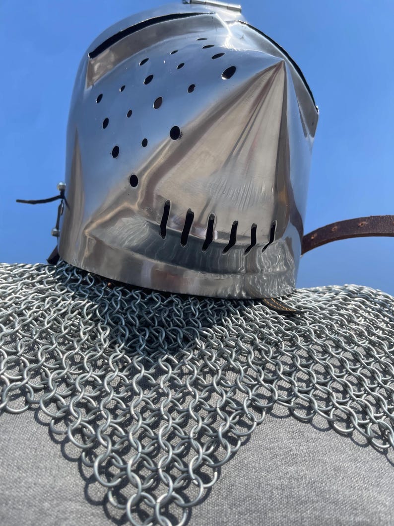 May include: A shiny, silver knight's helmet with a pointed visor and ventilation holes. The helmet rests on a chainmail coif, with a leather strap. The background is a clear, bright blue sky.