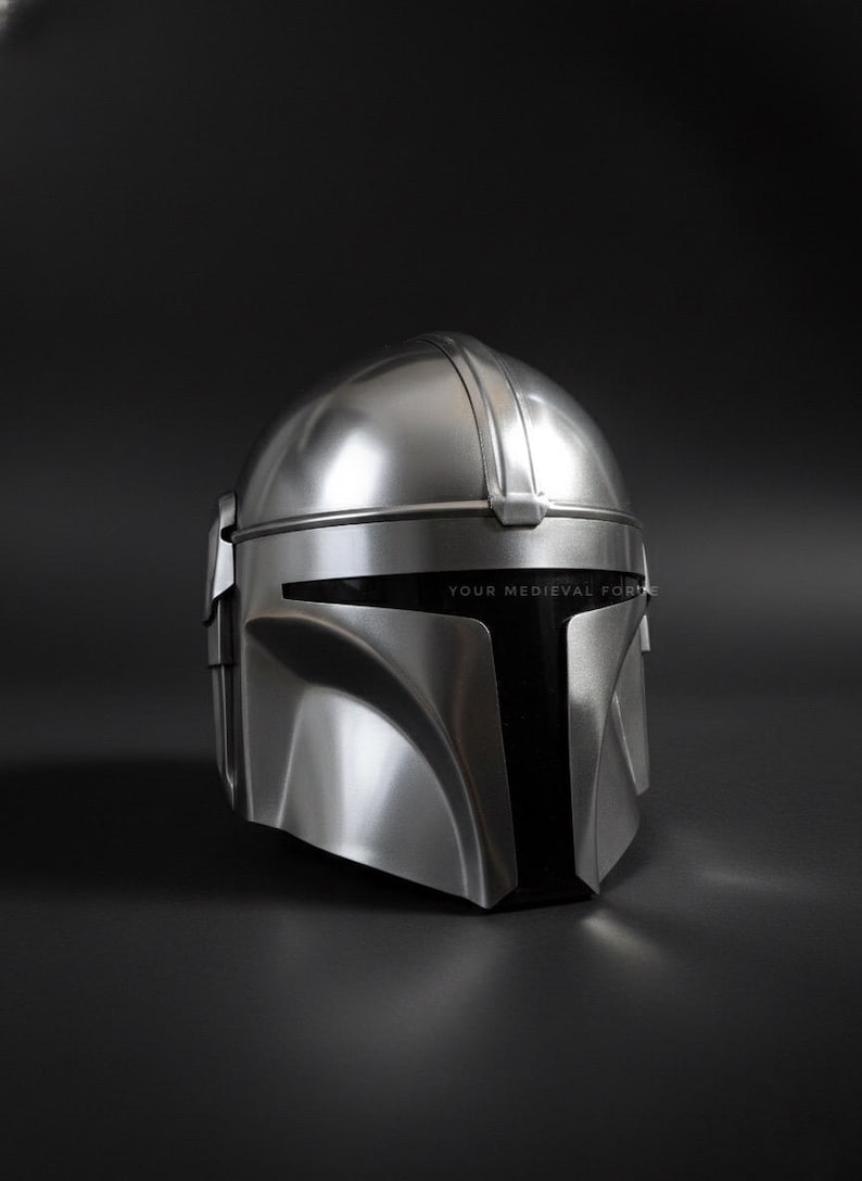 May include: A silver-colored helmet with a sleek, futuristic design. The helmet has a rounded top with a central ridge and a visor area. The text "YOUR MEDIEVAL FOR" is printed on the visor. The helmet is set against a dark background.