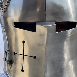 Peut inclure: Gros plan sur un casque de chevalier argent&eacute; et poli. Le casque pr&eacute;sente une croix sur le devant et des fentes pour les yeux. Une cha&icirc;ne est attach&eacute;e sur le c&ocirc;t&eacute;. Le casque est en m&eacute;tal.