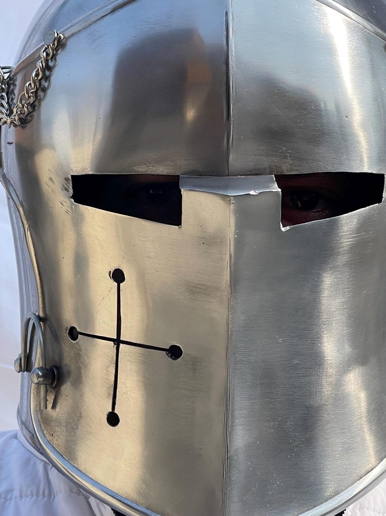 Peut inclure: Gros plan sur un casque de chevalier argent&eacute; et poli. Le casque pr&eacute;sente une croix sur le devant et des fentes pour les yeux. Une cha&icirc;ne est attach&eacute;e sur le c&ocirc;t&eacute;. Le casque est en m&eacute;tal.