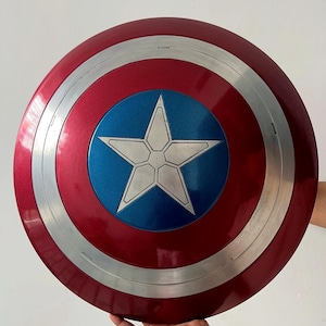 May include: A red, white, and blue Captain America shield. The shield has a metallic finish with a silver band and a blue center featuring a white star. The shield is round and appears to be a replica.