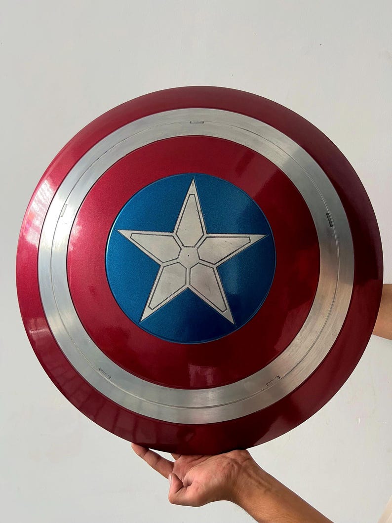 May include: A red, white, and blue Captain America shield. The shield has a metallic finish with a silver band and a blue center featuring a white star. The shield is round and appears to be a replica.