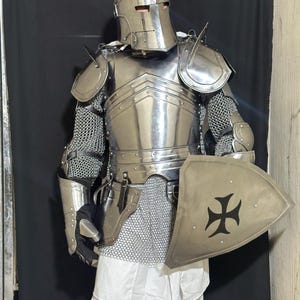 May include: A full suit of medieval armor, including a helmet, chest plate, shoulder guards, and chainmail. The armor is silver-colored and includes a shield with a black cross design. The person is wearing a white tunic.