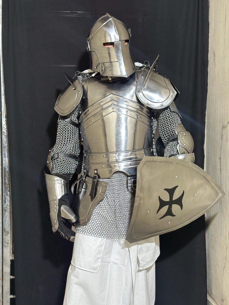 May include: A full suit of medieval armor, including a helmet, chest plate, shoulder guards, and chainmail. The armor is silver-colored and includes a shield with a black cross design. The person is wearing a white tunic.