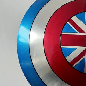 May include: A metallic blue, silver, and red shield with a Union Jack design. The shield has a circular shape with concentric rings of color. The blue ring is on the outside, followed by silver, red, and a central design.