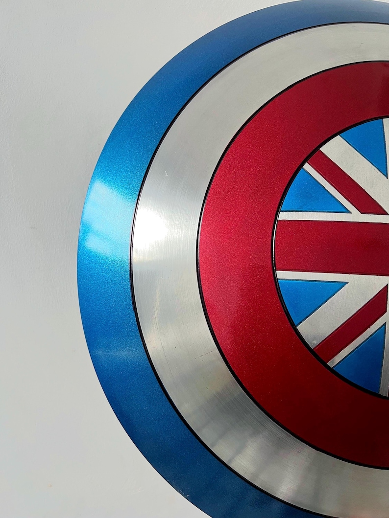 May include: A metallic blue, silver, and red shield with a Union Jack design. The shield has a circular shape with concentric rings of color. The blue ring is on the outside, followed by silver, red, and a central design.