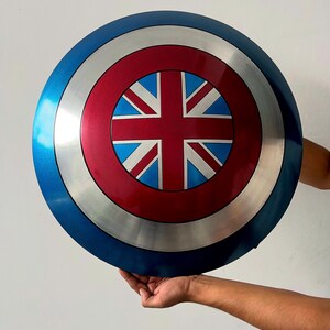 May include: A round shield with a blue, silver, and red color scheme. The center features a red circle with a Union Jack design. The shield is held up by a person's hands.