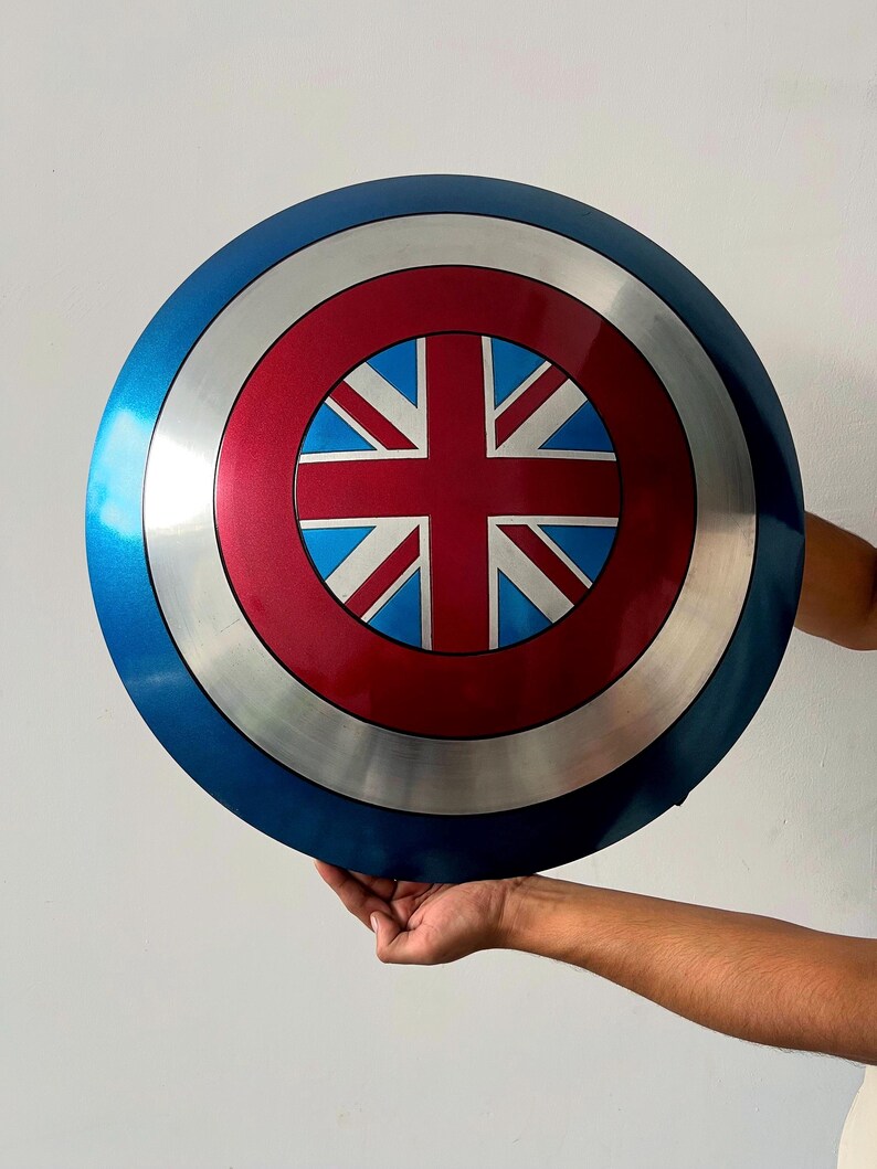 May include: A round shield with a blue, silver, and red color scheme. The center features a red circle with a Union Jack design. The shield is held up by a person's hands.
