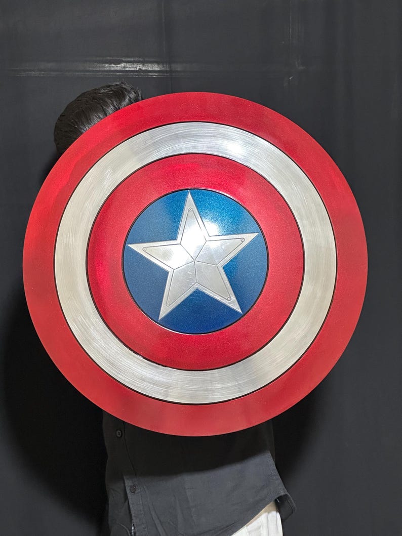 May include: A red, white, and blue shield with a silver star in the center. The shield has a circular design with concentric rings of red, silver, and white. The center is blue with a silver star.