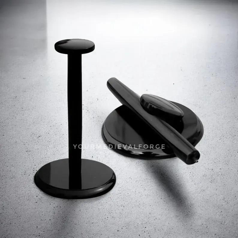 May include: Two black metal display stands. One is a tall, cylindrical stand with a circular base and a flat top. The other is a shorter, circular stand with a curved handle and a small oval-shaped piece. The stands are on a light gray surface.