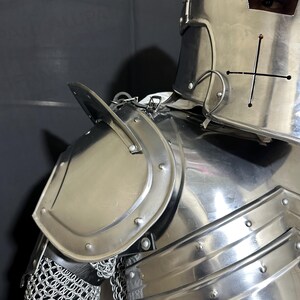 May include: Close-up of a polished, silver-colored knight's armor. The armor includes a helmet with a cross design, shoulder plates, and a chainmail section. The image highlights the metallic sheen and intricate details of the medieval-style protective gear.