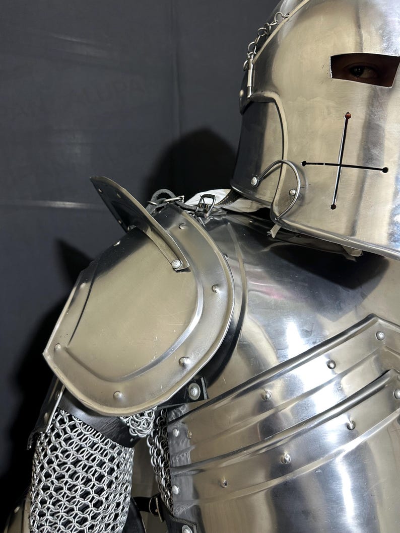 May include: Close-up of a polished, silver-colored knight's armor. The armor includes a helmet with a cross design, shoulder plates, and a chainmail section. The image highlights the metallic sheen and intricate details of the medieval-style protective gear.