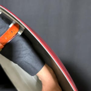 May include: A red and silver shield with a red border. The shield has a silver interior and a red outer edge. A brown leather strap is visible. The shield is held by a person.