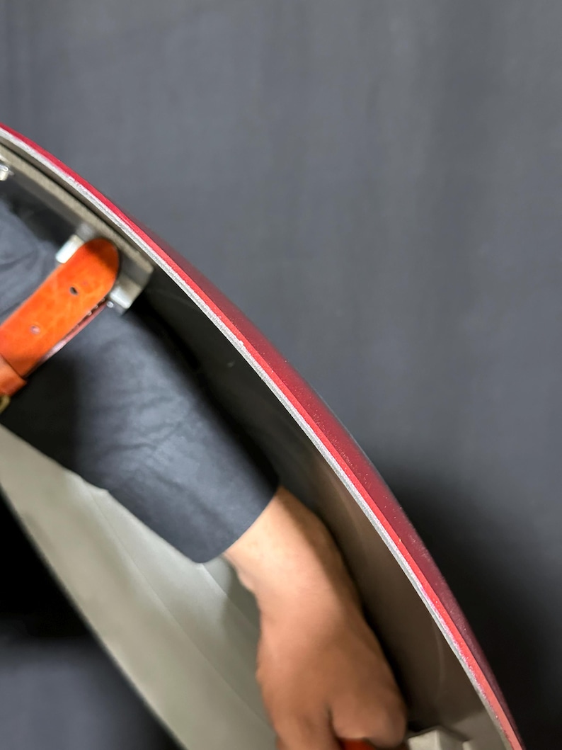 May include: A red and silver shield with a red border. The shield has a silver interior and a red outer edge. A brown leather strap is visible. The shield is held by a person.