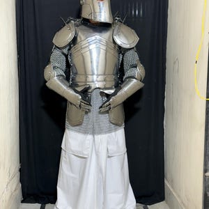 May include: A full suit of polished steel armor, including a helmet, chest plate, shoulder guards, and arm protection. The armor is worn over chainmail and white pants. The person is wearing sandals.