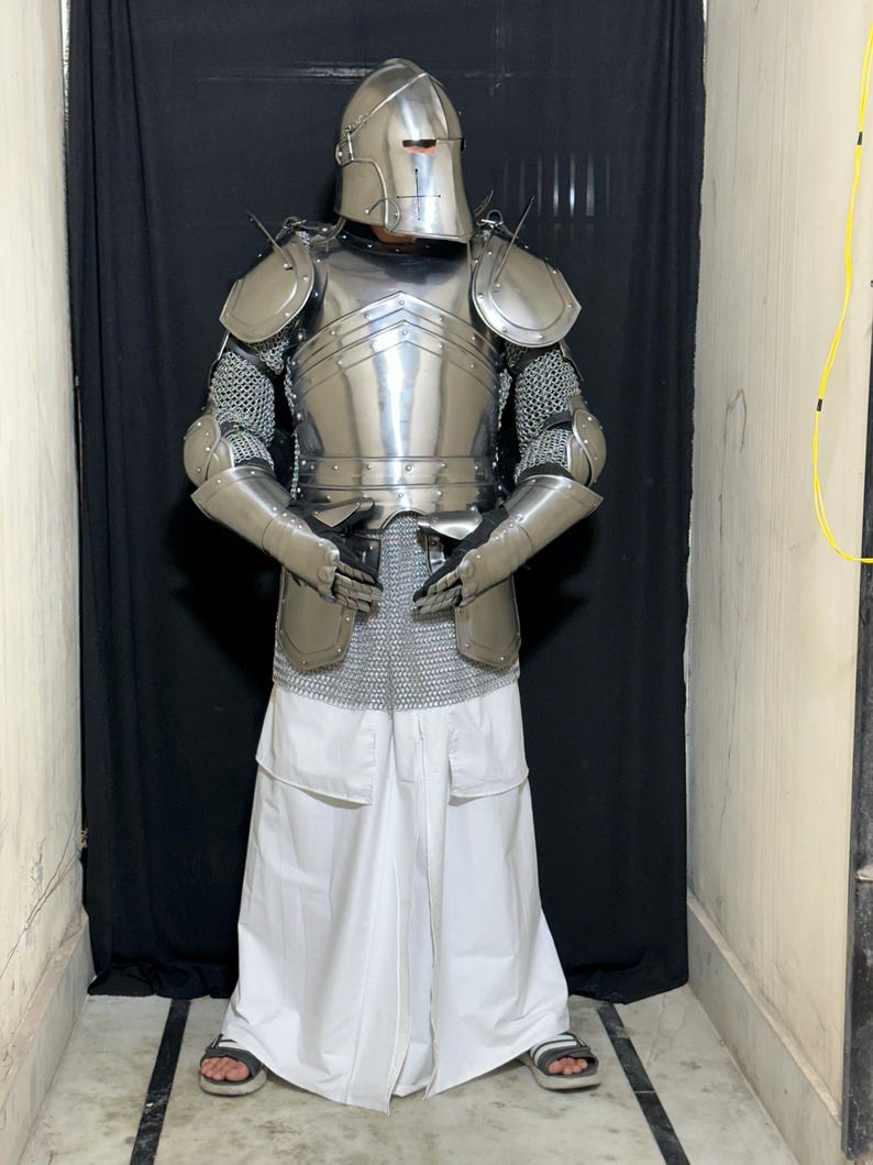 May include: A full suit of polished steel armor, including a helmet, chest plate, shoulder guards, and arm protection. The armor is worn over chainmail and white pants. The person is wearing sandals.