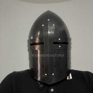 May include: A black medieval helmet with a rounded top and a visor with horizontal eye slits. The helmet has rivets and a cross-shaped reinforcement. The text "YOURMEDIEVALFORGE" is visible on the front of the helmet.
