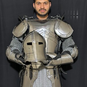 May include: A full suit of polished steel armor, including a helmet, chest plate, shoulder guards, and arm protection. The armor is connected with chainmail. The helmet has a cross design. The armor is designed for protection.
