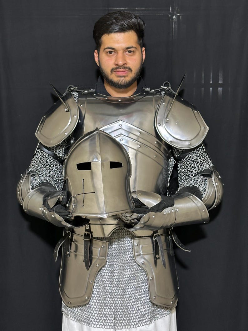May include: A full suit of polished steel armor, including a helmet, chest plate, shoulder guards, and arm protection. The armor is connected with chainmail. The helmet has a cross design. The armor is designed for protection.
